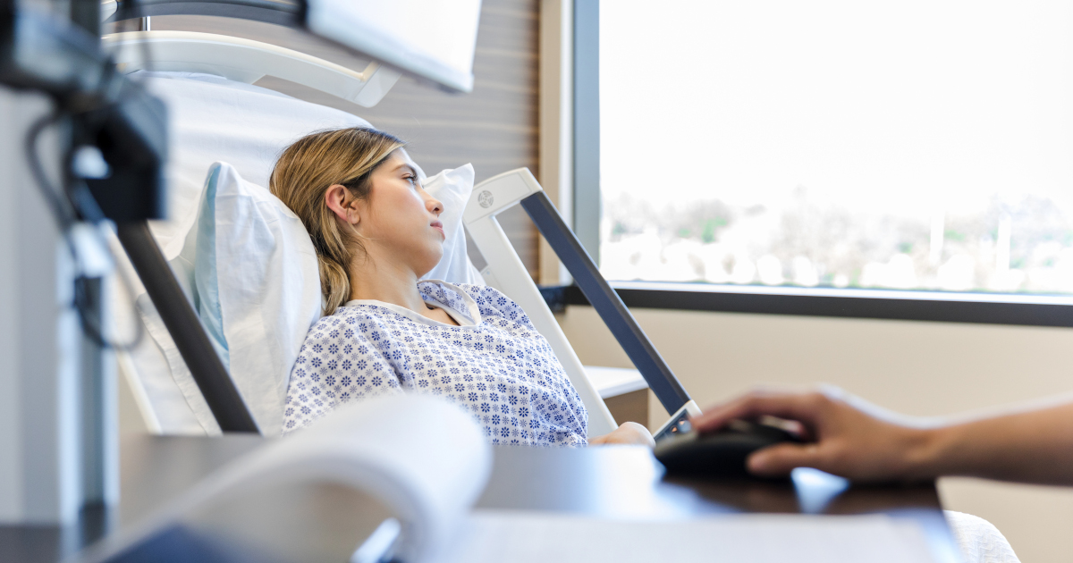 A Young Woman In A Hosptial Gown Lies In A Hospital Bed Looking Out The Window In The Daytime, While In The Foregound A Hand Works A Computer Mouse In Front Of A Monitor