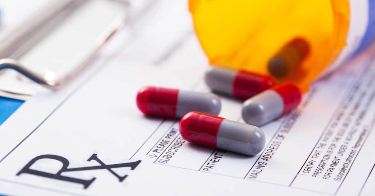 An Orange Pill Bottle With The Cap Off and Several Red And Gray Pills Lying Atop a Pharmacy Perscription Form