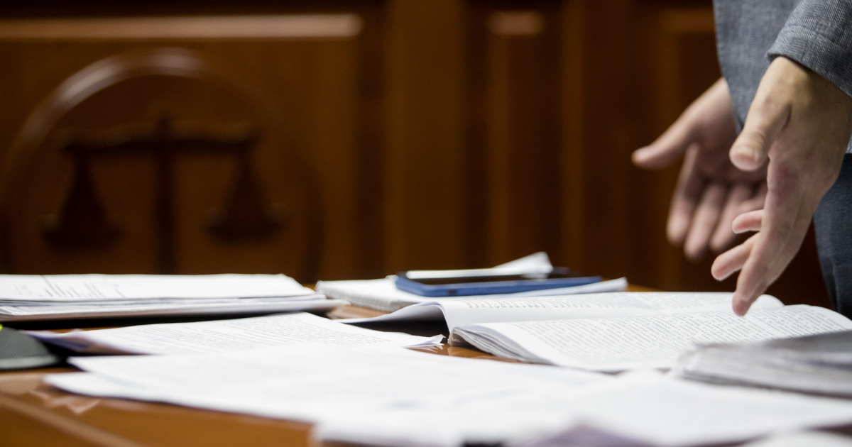 View of An Attorney In A Dark Suit, Seen From The Elbows Down, Gesturing With His Palms Up Above Papers Strewn Across Counsel's Table In A Courtroom