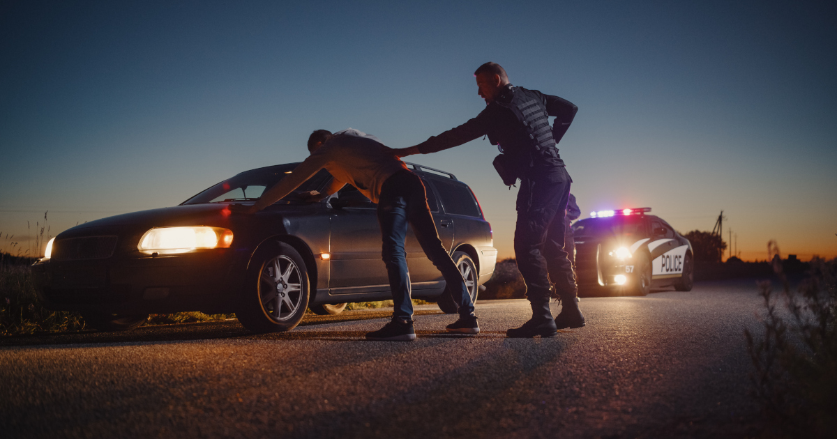 Backlit By A Summer Sunset, A Police Man Pats Down A Driver Who Is Spreadealged Against The Side Of A Station Wagon