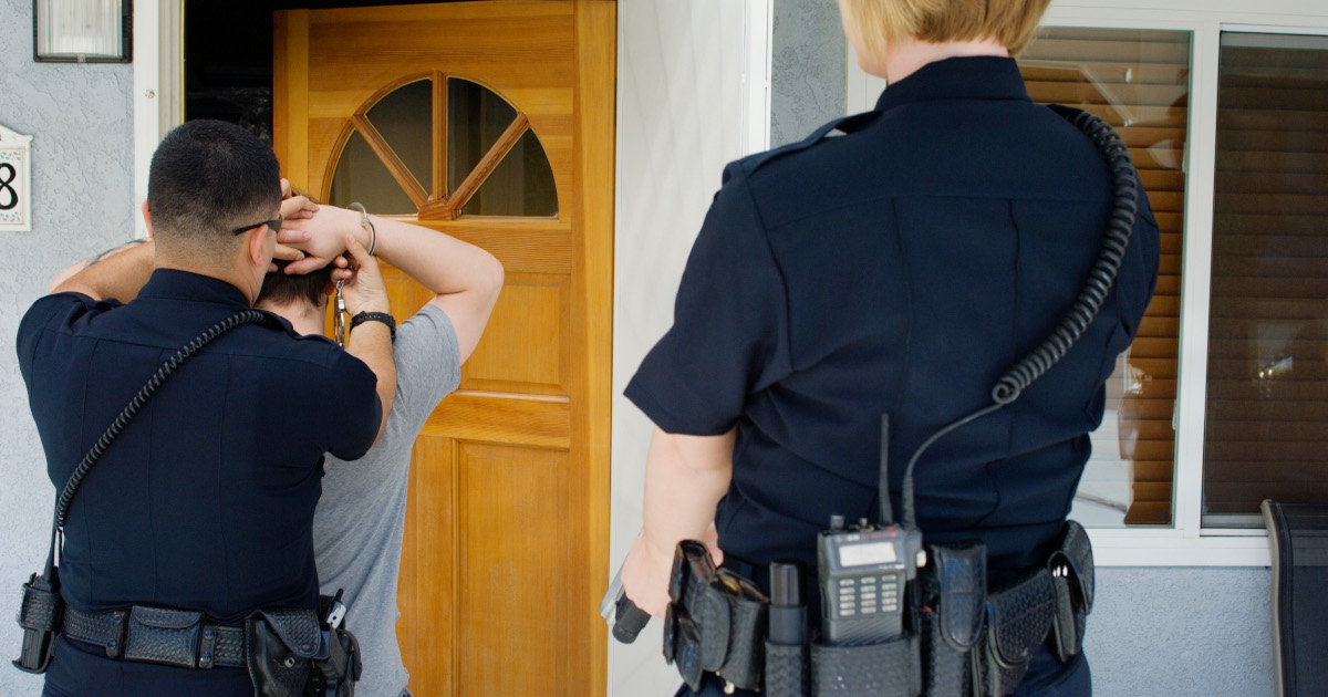 Two Police Officers, One A Man, The Other A Woman, Seen From Behind As The Male Officer Handcuffs A Man On His Front Porch, With The Front Door Half Open In The Background