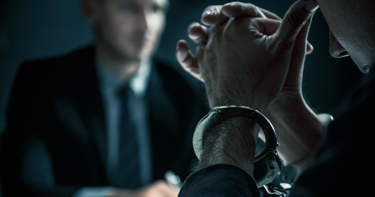 In The Blue Gray Gloom Of A Police Interrogation Room, A Handcuffed Suspect In The Foreground Rests His Forehead On Steepeld Thumbs, While A Detective In The Background Sits With A Pen Poised Over A Piece Of Paper