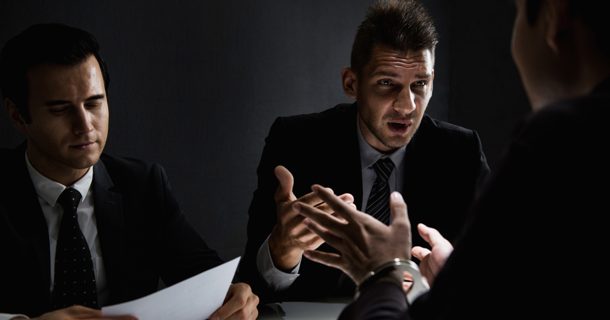 In A Half-Lit Room,Two White Men Wearing Dark Suits And Ties Sit Gesturing Across A Table From A Handcuffed Man Wearing A Dark Shirt