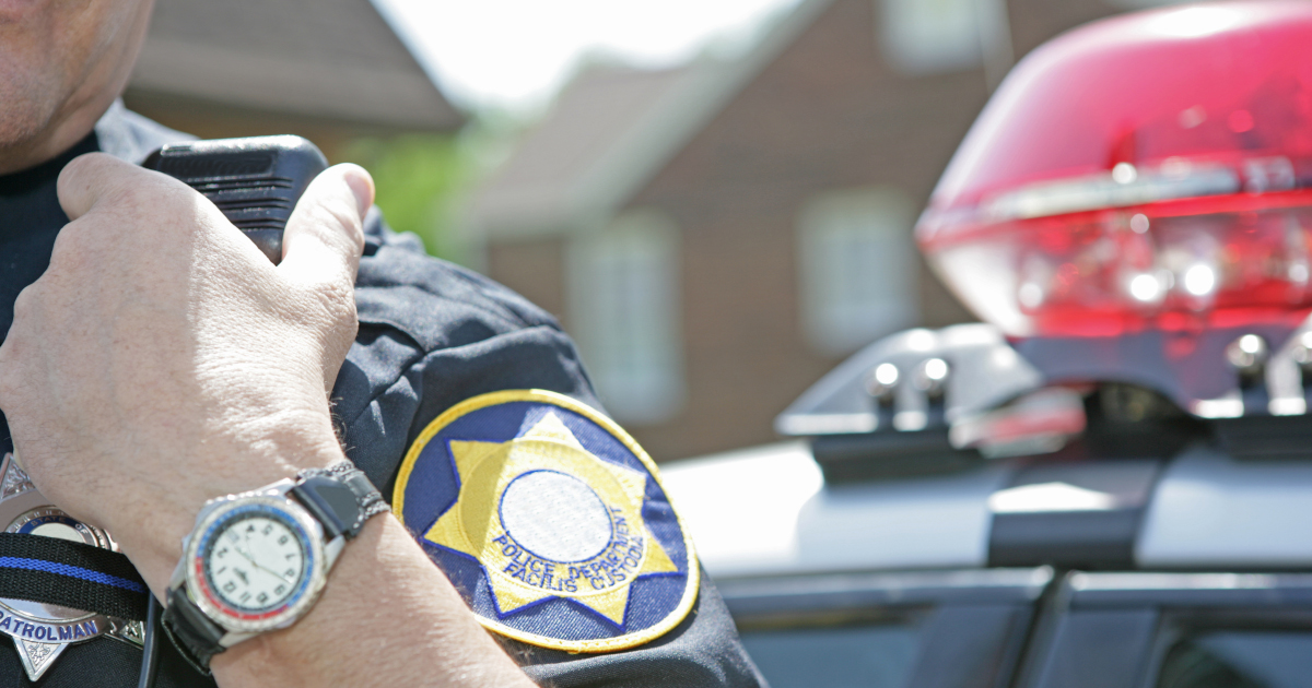 As He Stands In Front Of A House, A Policeman Reaches To Activate The Radio Microphone Clipped To His Uniform