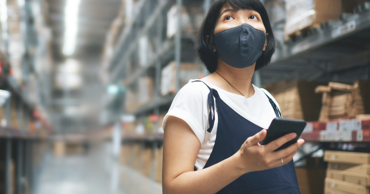 A Pregnant Worker Stands In A Warehouse And Looks Pensively Up At The Shelves While Talking On A Smart Phone