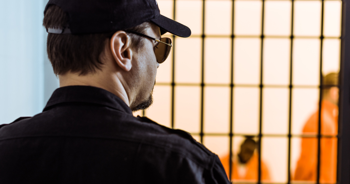 A Male Prison Guard In A Ball Cap And Aviator Shades Stands In the Foreground, With A Cell And Orange-clad Inmates Out of Focus In The Background