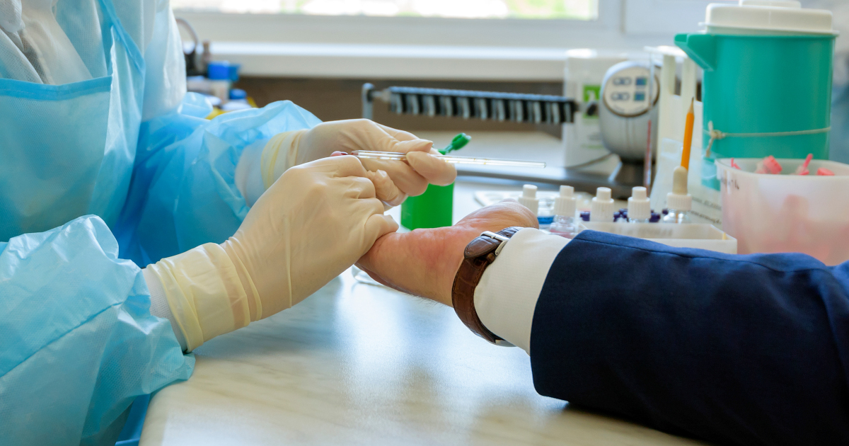 A Health Care Worker In Blue Scrubs And A Blue Smock And Latex Gloves Preparing to Jab a Man's Hand In A Laboratory