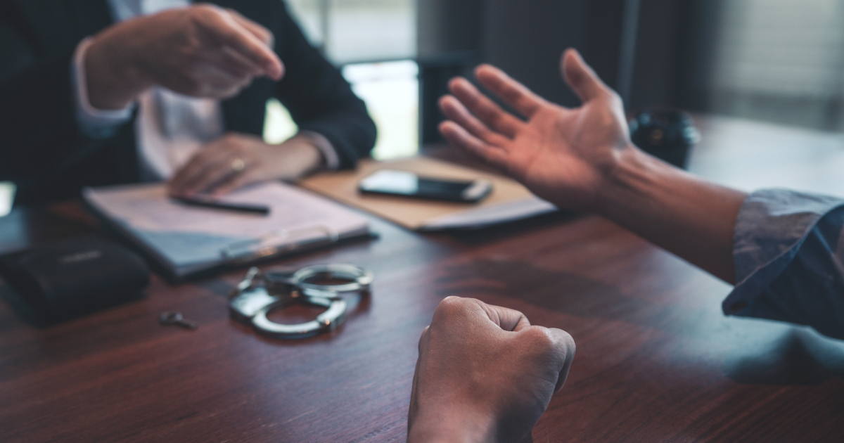 In A Poorly Lit Room A Man In A Suit Points Across A Table At Someone With Their Hand Extended Imploringly, With A Pair of Handcuffs On The Table Between Them,