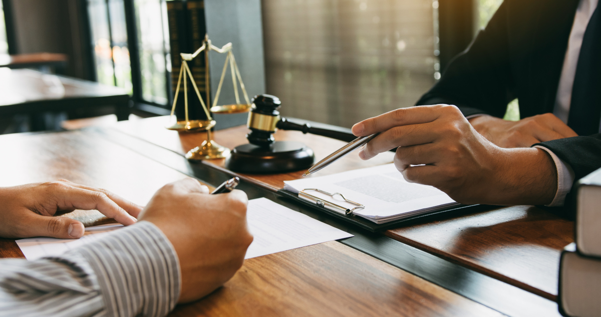 A Man In Shirtsleeves, Sitting Across A Desk From A Man In A Suit And Tie, Signs A Piece of Paper While The Man In The Suit Points At The Paper With His Pen, In The Background Window Light Illuminates A Scales Of Justice And A Judge’s Gavel