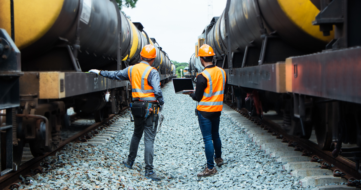 Two Male Workers, Clad In Orange HardHats and Yellow High Viz Vests, Stand Between A Row of Railroad Cars With Their Backs To The Camrea, With The Cars Stretching Away Into The Distance