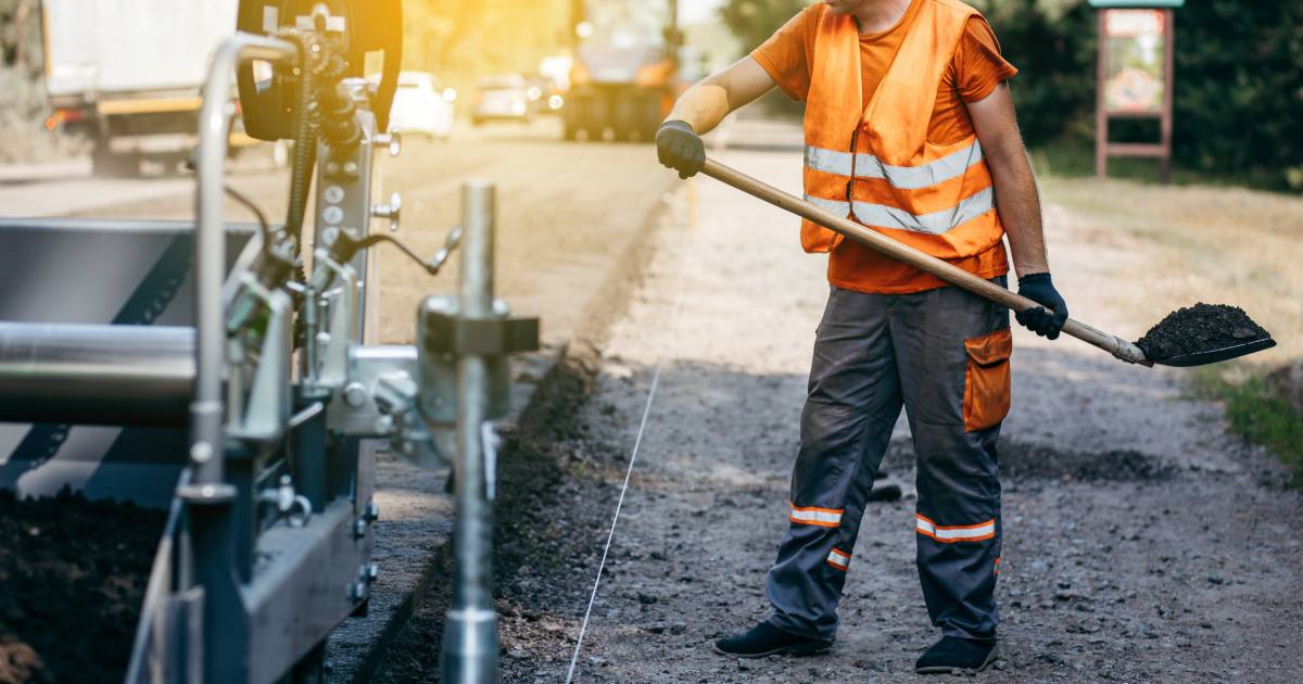 A Construction Worker In A Orange High Viz Vest Shovels Hot Asphalt Along The Shoulder Of A Freshly Black Topped Road