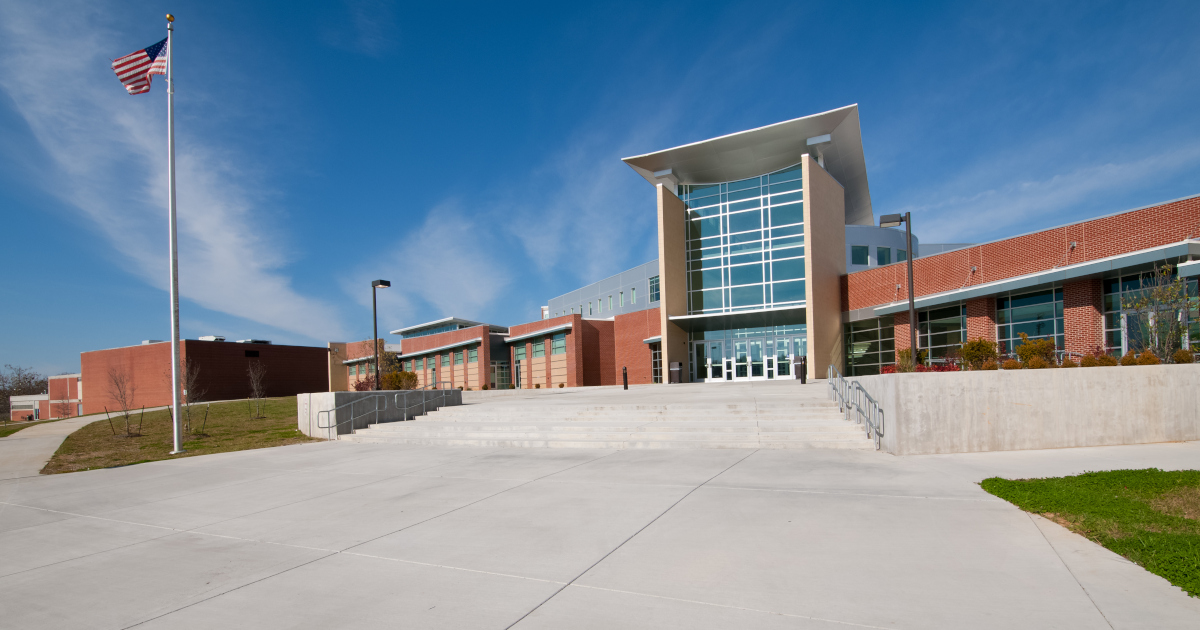 A Large, New Brick School Building Beneath A Blue Sky, With A Flagpole Out Front