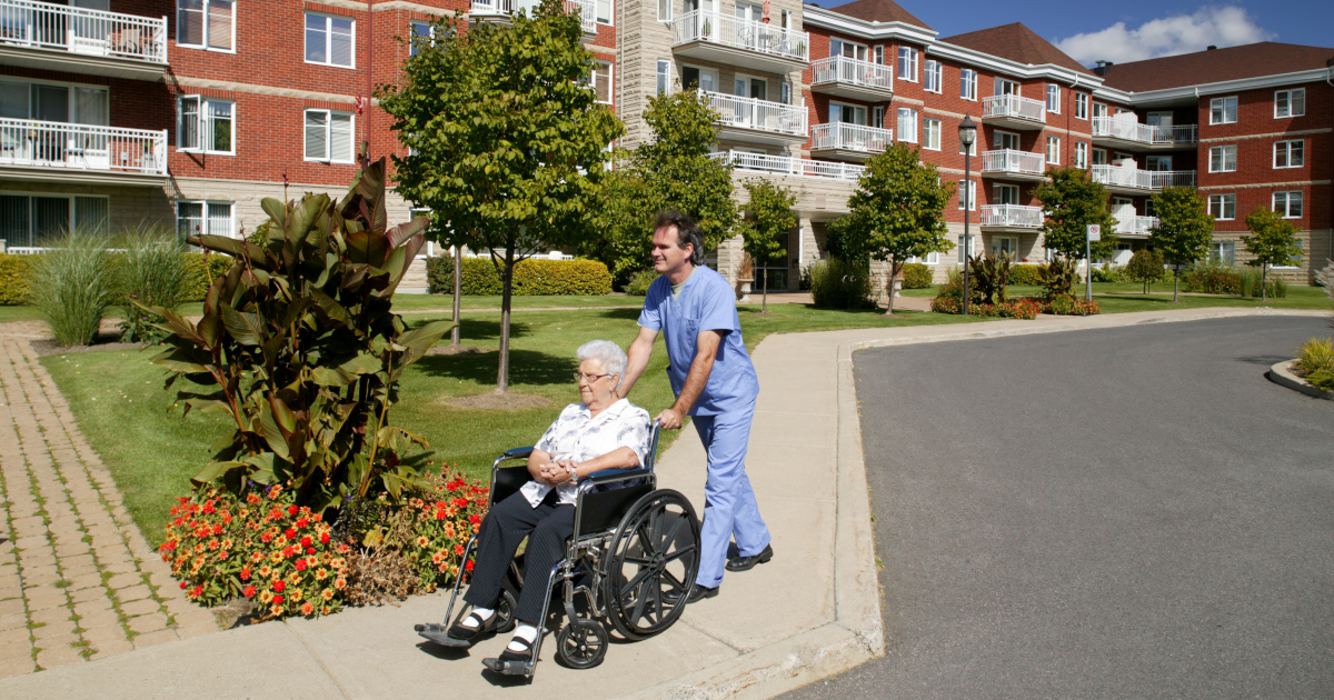 A Young Man In Blue Medical Smock And Pants Pushes A White-Haired Woman In A  Wheelchair Outside A Three-Story Housing Complex