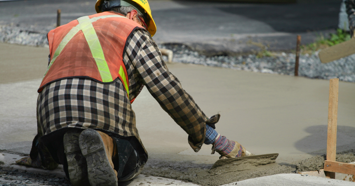 A Man In An Orange Safety Vest, A Yellow Hard Hat, And Work Boots Kneels On A Curb As He Uses A Trowel To Smooth Out Wet Concrete Poured To Make A Sidewalk