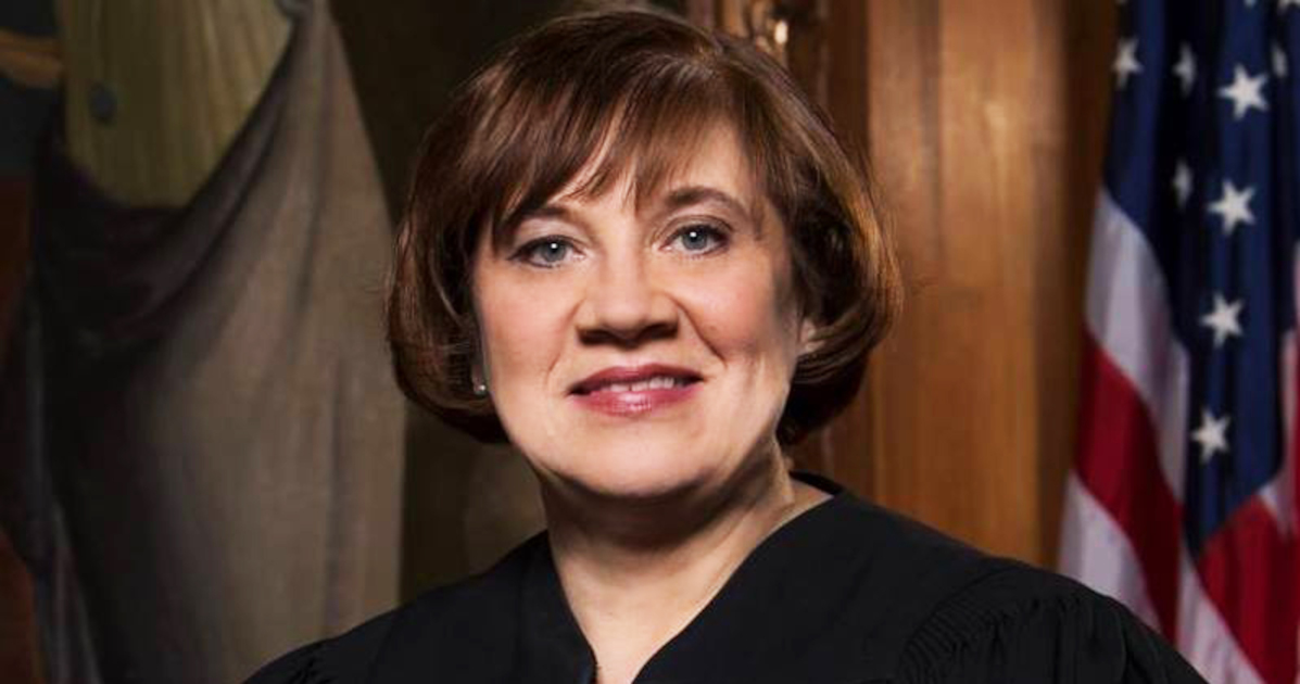 A Middle-Aged White Woman With A Brunette Bob In A Black Judge's Robe With The American Flag Over Her Left Shoulder