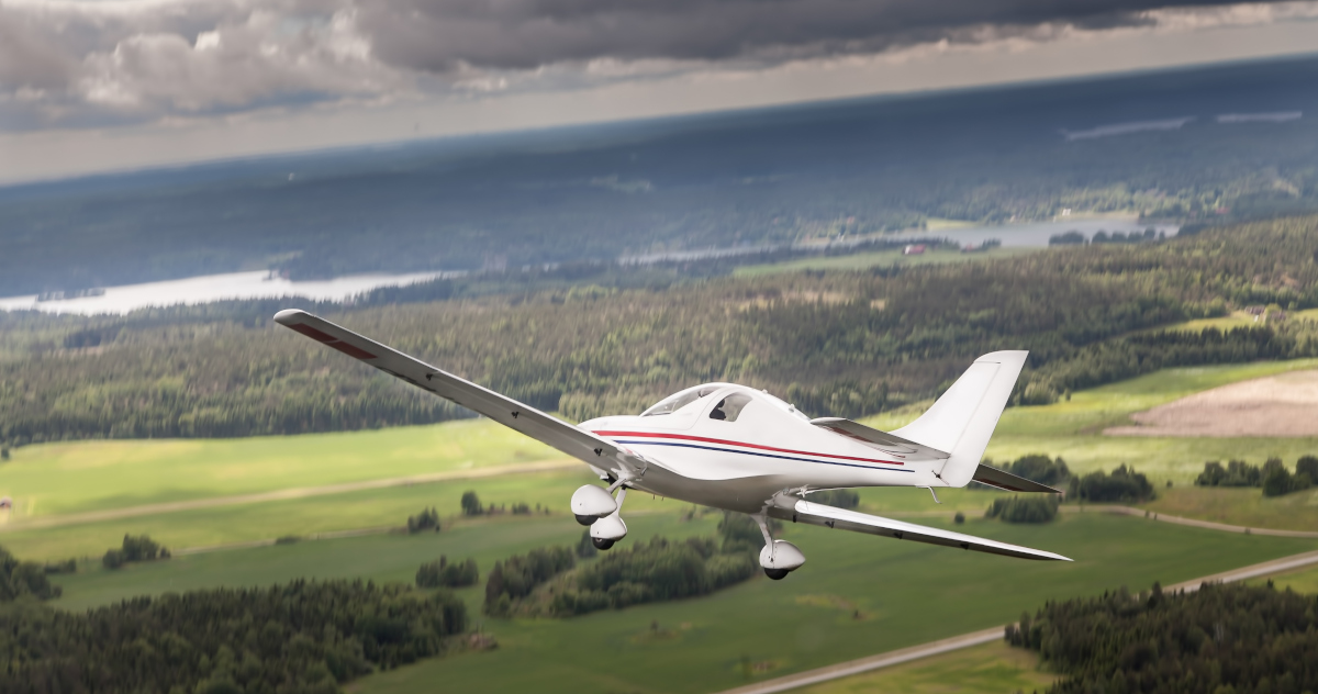 Beneath A Bank of Dark Gray Clouds, A Small, White Single-Engine Airplane Banks To The Right Above A Landscape Marked By Deciduous Trees And A Lake