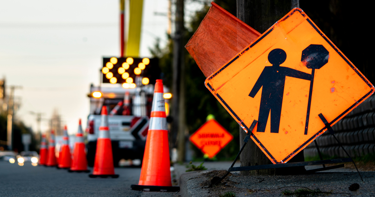 An Orange Roadside Sign Warning Of Workers Ahead, With Orange Construction Cones Stretching Along The Roadside Into The Distance