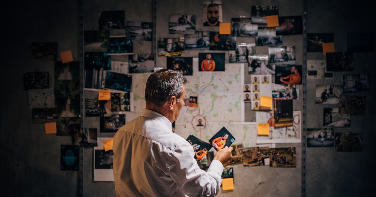 Standing With His Back To The Camrea, A Middle-Aged Detective Pins A Photo Of A Suspect To A Bulletin Board Filled With Photos Of Other Suspects