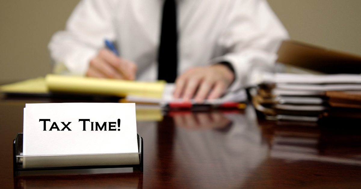 An Man In A White Dress Shirt And A Black Tie Sits Behind A Table Full of Forms, With A Small Sign That Reads Tax Time In The Foreground