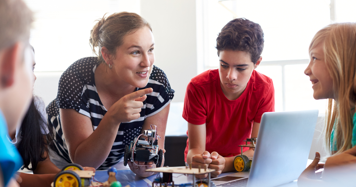 A Teacher Leaning Over A Work Table, Gesturing And Talking To Smiling Students