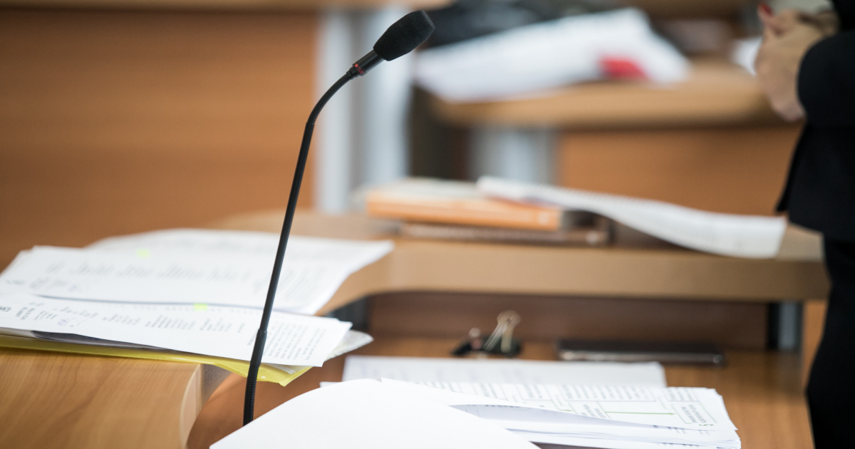 Stacks Of Legal Papers Heaped Upon A Table In A Courtroom While A Lawyer Stands Behind The Table