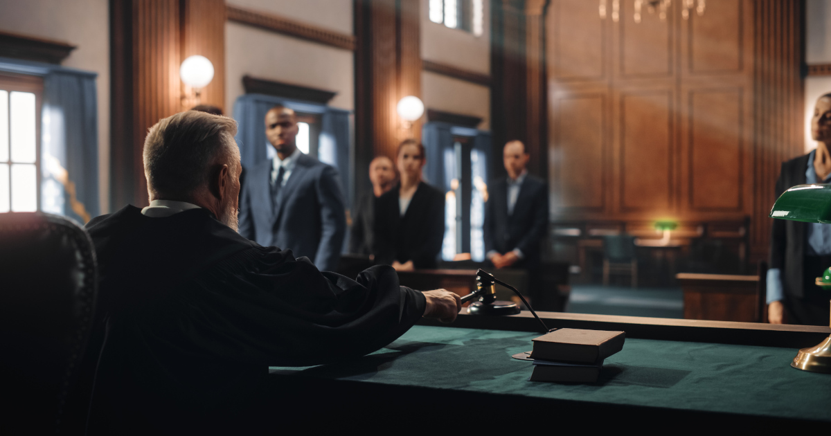 Two Men and Two Women, Dressed In Dark Business Suits, Standing Behind A Counsel Table As A Judge Gavels A Proceeding To Order