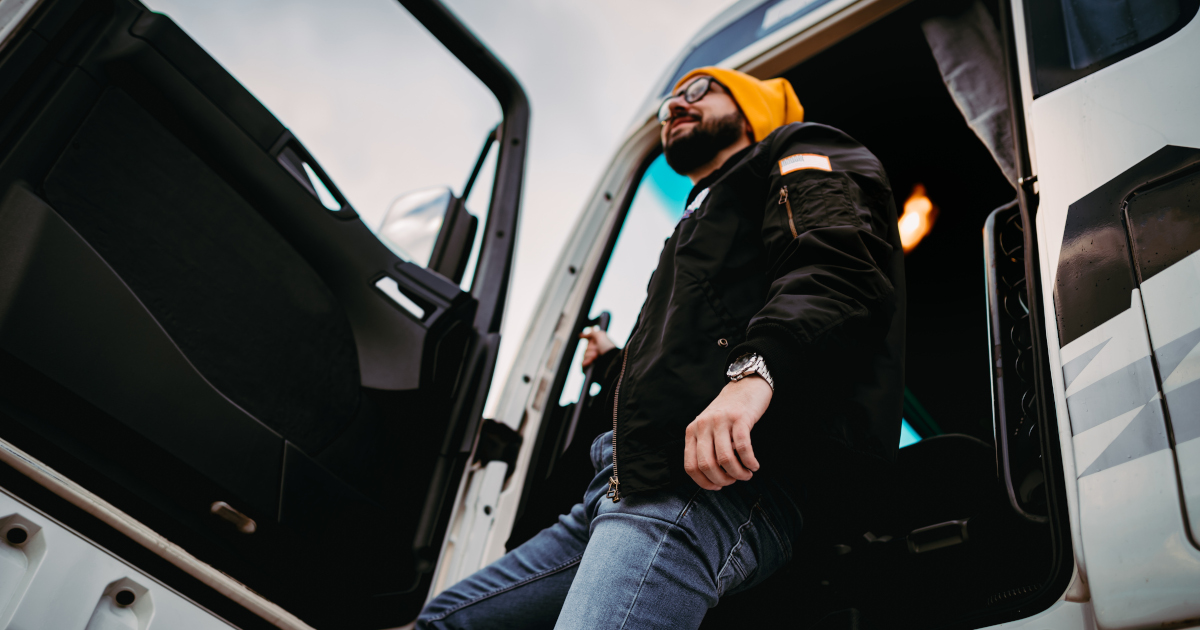 A Bearded Man, Clad in Jeans And A Mesh Ball Cap, Stepping Down From The Cab Of A Tractor-Trailer Cab