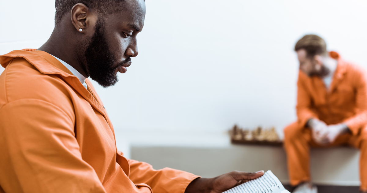 In The Foreground, A Prisoner In An Orange Jumpsuit Sits Reading A Book; In The Background Another Prisoner In An Orange Jumpsuit Sits On A Bench Looking Down At A Chess Board