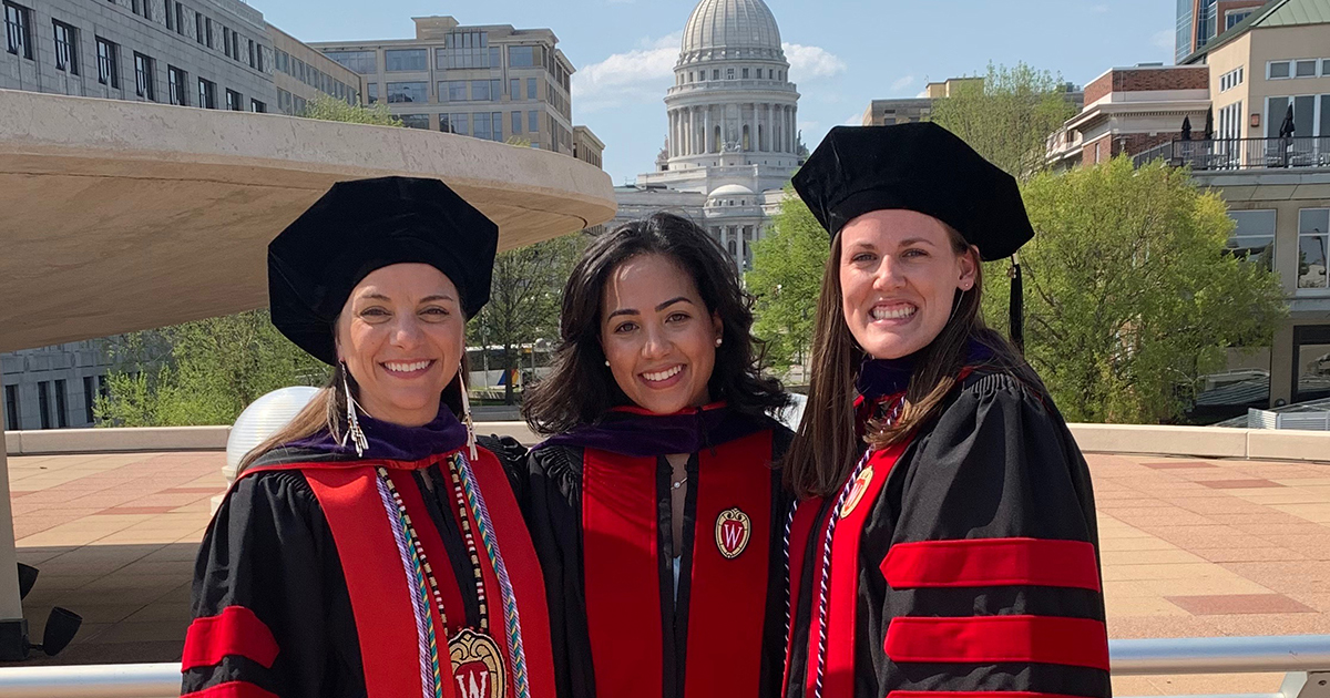 three women in academic regalia stand with the Capitol in the background