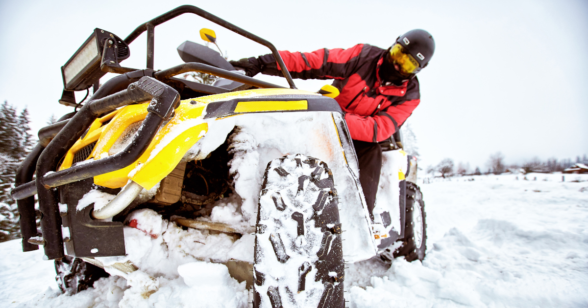 A Wide Angled Closeup, Shot From The Ground, Of A Man In A Red Snowsuit And A Black Helmet On Yellow Four-Wheeled Utility Vehicle On The Snow