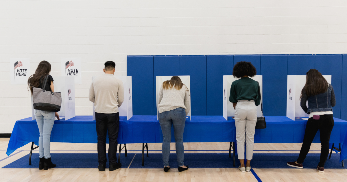 Four People In A Line Bent Over Their Ballots In Voting Bootsh, With Their Backs To The Camera