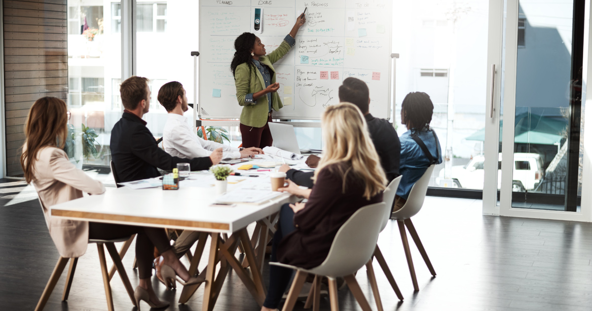 A Group Of Business People Sitting Around A Table While A Female Executive Stands At A Whiteboard And Points To Figures On The Board 