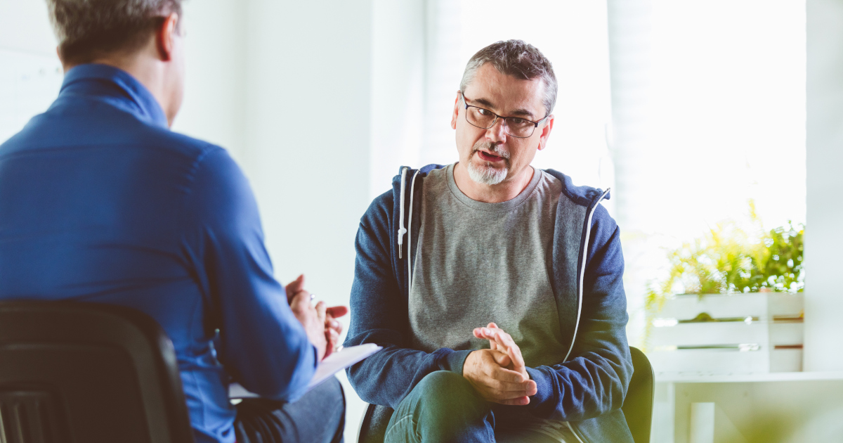 A Middle-Aged Man Dressed In A Hoodie, With His Hands Clasped, Sits In A Chair Facing A Therapist In A Brightly Lit Office