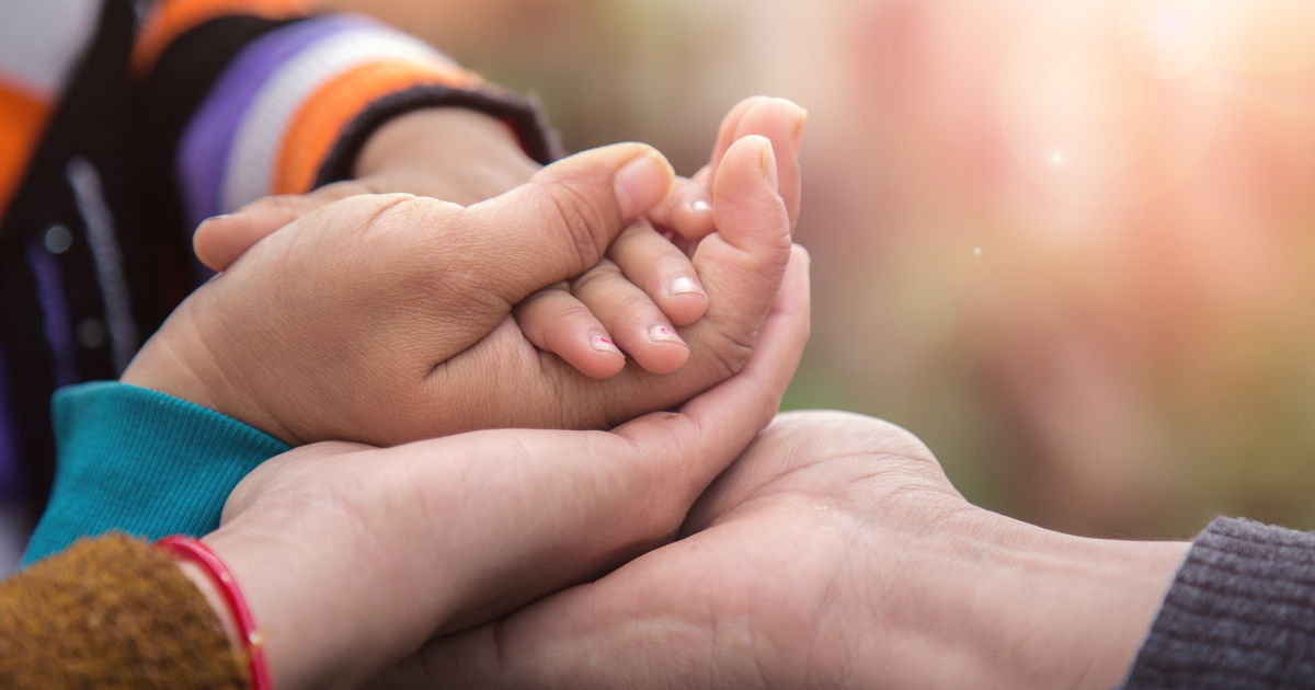A Closeup Of One Hand Each From Two Adults, Each Extended Palms Up And On Top Of Each Other, With A Child's Hand Placed Palm Down Atop The Adult's Hands, With The Background Artfully Blurred