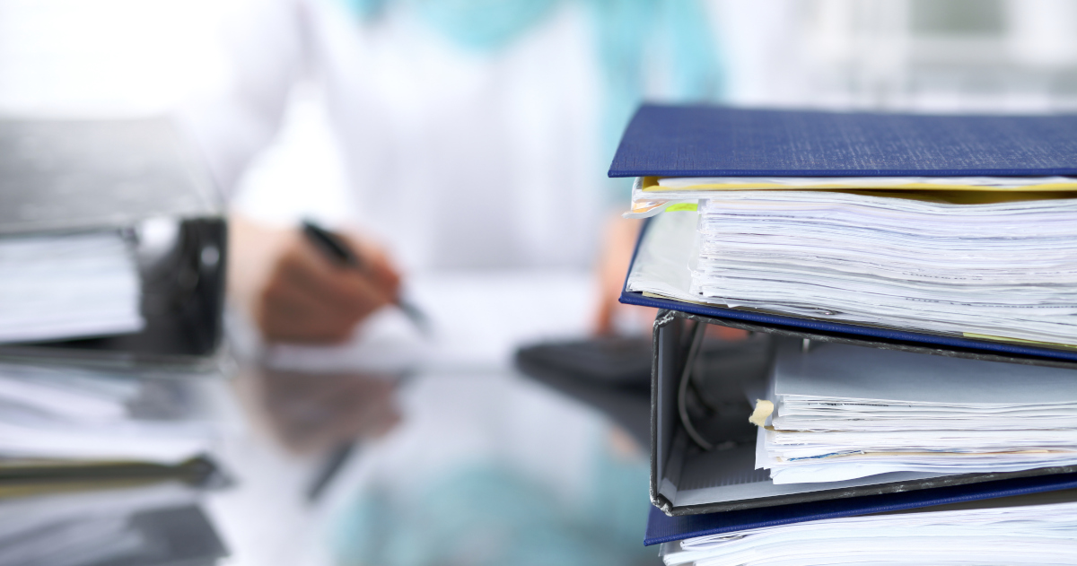 Blue Three-Ring Binders Full of Documents Stacked Atop A Desk While A Woman Writes In The Background