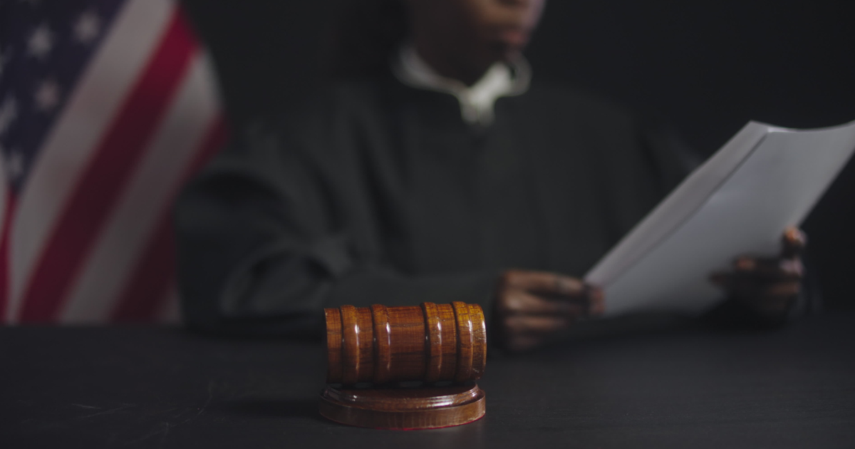 Medium Shot Of A Male Judge In A Black Robe, Slightly Out Of Focus, Seated On His Dais Reading A Document With THe American Flag In the Back Left Corner, With A Gavel In The Left Foreground