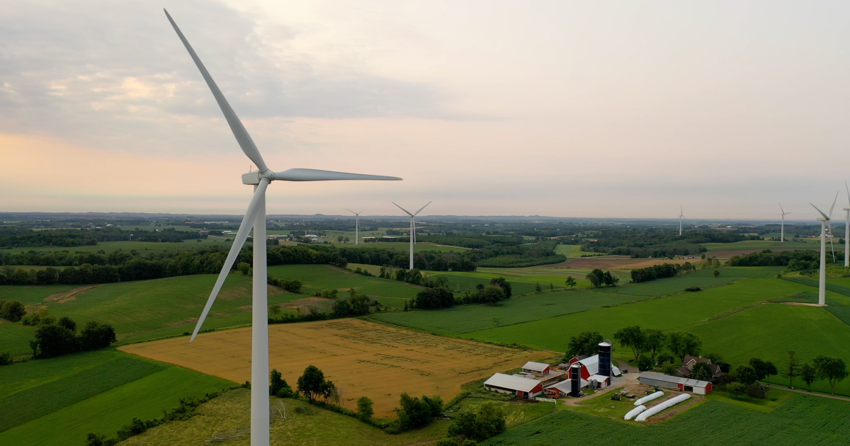 A Farm House, Barn, And Silos Set Down Amid Wind Turbines Stretching Off Into The Distance